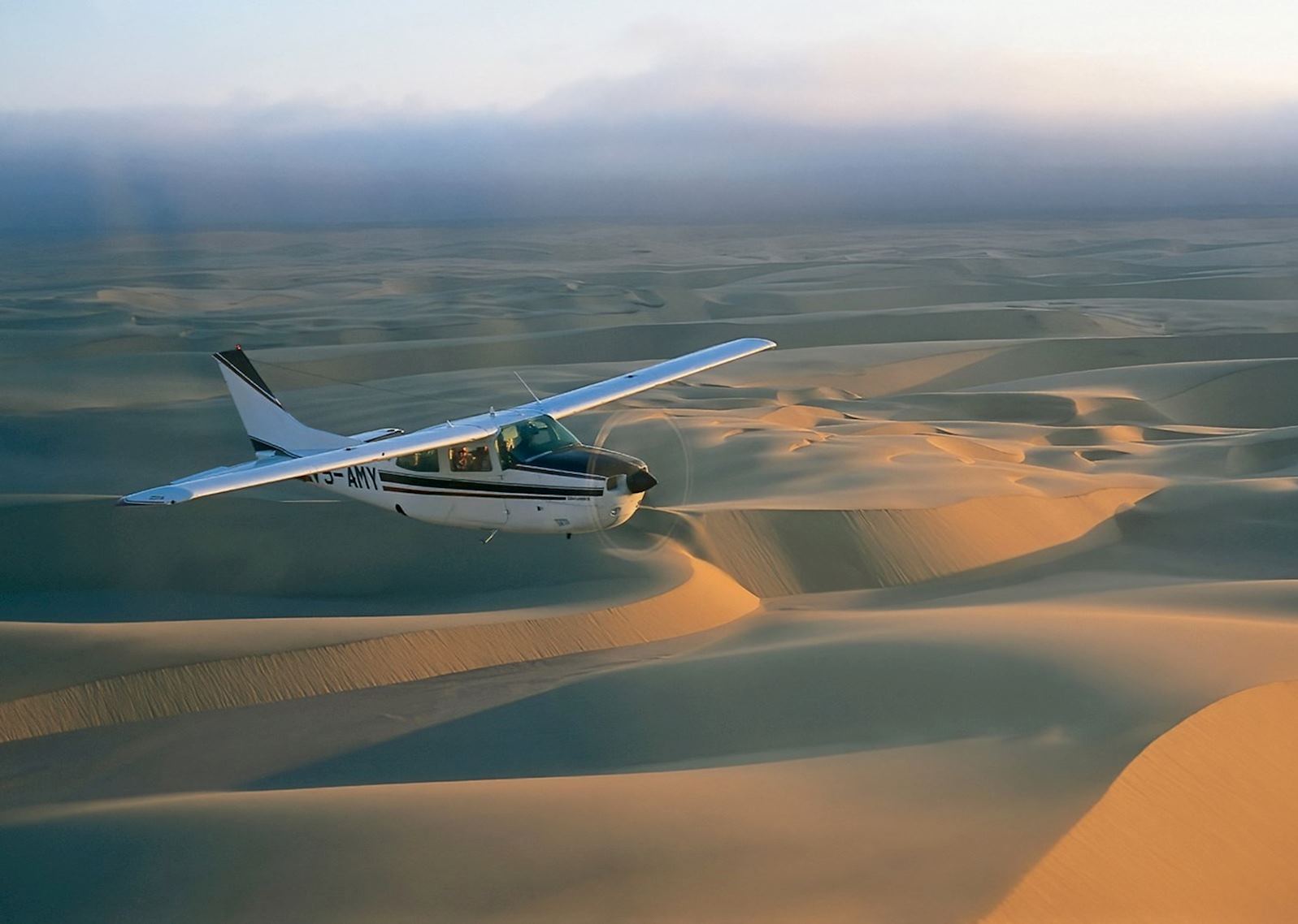 A small aircraft flying over vast desert dunes at sunrise, showcasing smooth, undulating sand patterns under a cloudy sky.