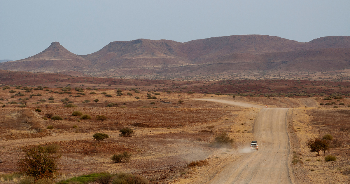 Car driving along a wide gravel road through hilly country, dust trailing behind, Namibia